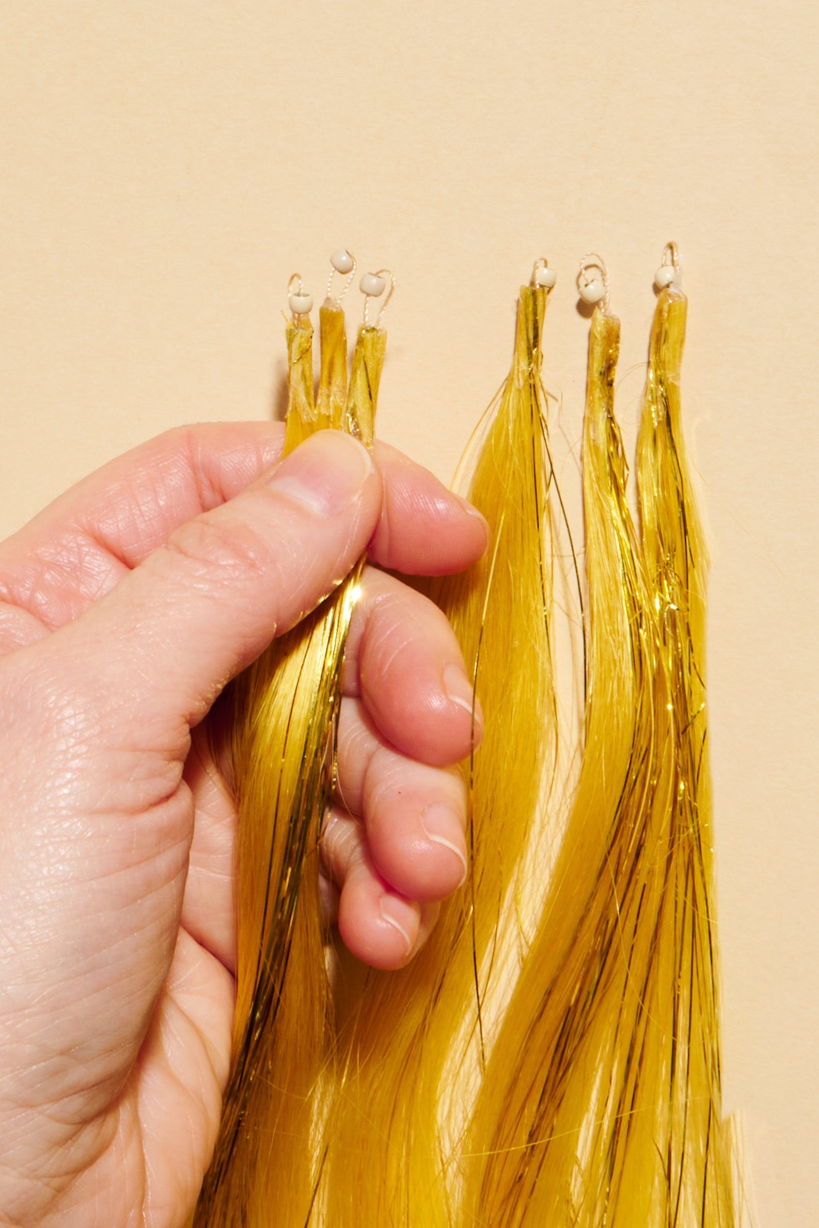 פקשוט של ידיים, המחזיקות תוספות שיער טבעיות, במרקם חלק - בצבע צהוב. בקצה התוספות שזורים חרוזי מעיכה בצבע בלונדיני